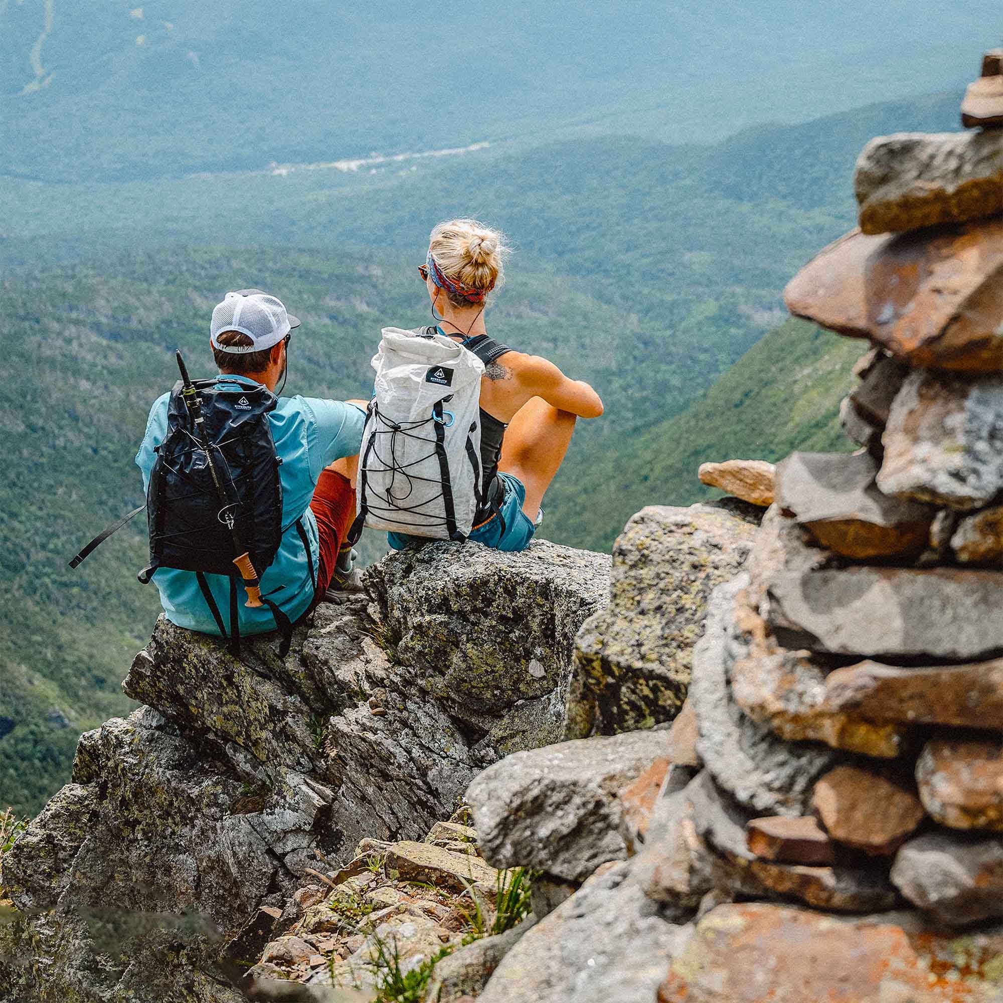Two people sitting on top of a rock overlooking a valley.